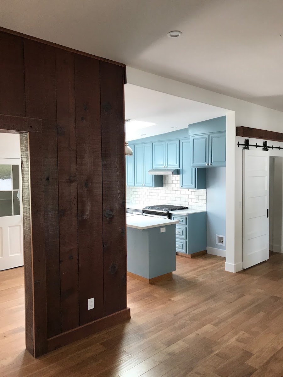 A finished open-plan interior. In the foreground a wall clad in dark
                  stained reclaimed boards frames a doorway; beyond it a kitchen in soft
                  blue shaker cabinets sits against a white subway tile backsplash, with
                  a matching island, a stainless range and a skylight above. A white
                  paneled barn door hangs on a black rail to the right, over wide oak
                  floorboards.