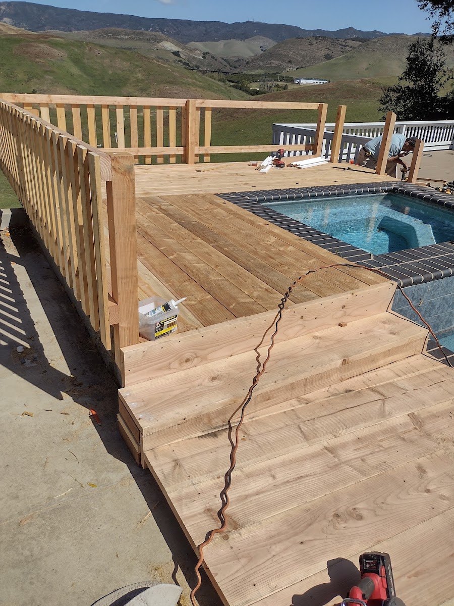 A new douglas fir deck being built beside a swimming pool. Fresh boards
                  run from the foreground to the water's edge, a wide step drops down to
                  a lower level, and a slatted railing is going up along the left side. A
                  man works on the far rail beyond the pool, with green Central Coast
                  hills rising behind the property.